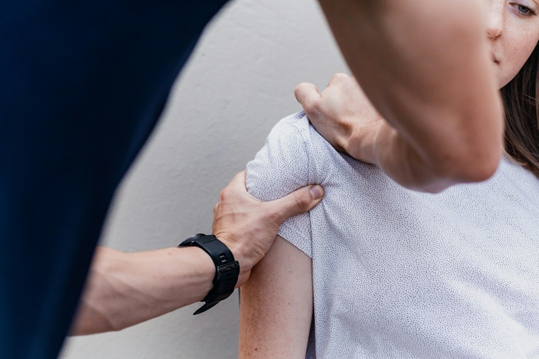 A person massaging the shoulder of another person wearing a white patterned shirt